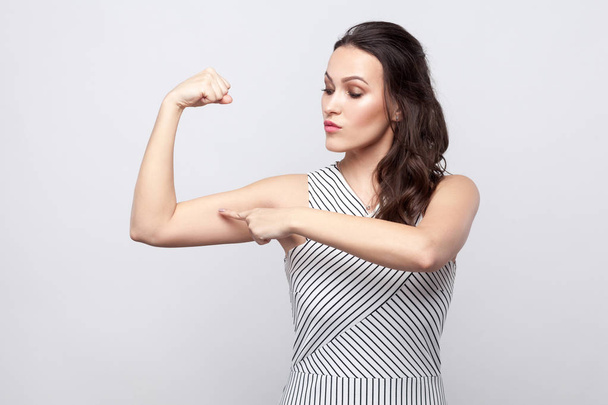 Portrait of proud beautiful young brunette woman with makeup and striped dress standing and pointing at her biceps on grey background. - Фото, зображення