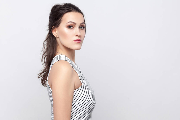 Profile side view portrait of serious beautiful young brunette woman with makeup and striped dress standing and looking at camera with serious face on grey background. - Photo, image