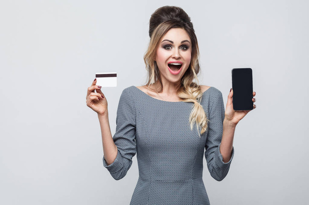 Portrait of happy beautiful modern woman in grey dress holding smartphone and credit card with open mouth and looking at camera on grey background  - Foto, imagen