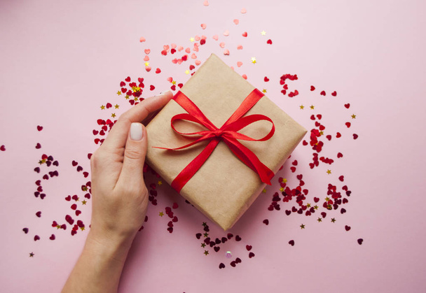 Gift box wrapped in brown colored craft paper and tied with red bow on pink background with red confetti. Lady's hand holding a giftbox. - Fotoğraf, Görsel
