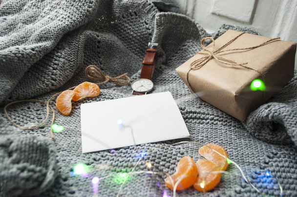 Теплые выходные в холодную погоду. Подарочная коробка, часы, мандарин и кусок бумаги на сером вязаном фоне с гирляндой
. - Фото, изображение