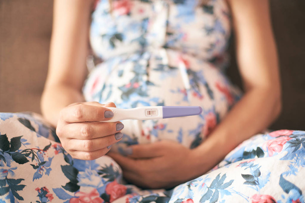 Gros plan du test de grossesse dans les mains de la jeune femme en robe assise sur le canapé à la maison
 - Photo, image