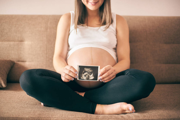 terhes nő ül a kanapén, otthon és találat ultrahang kép a gyermek nagy hasa  - Fotó, kép