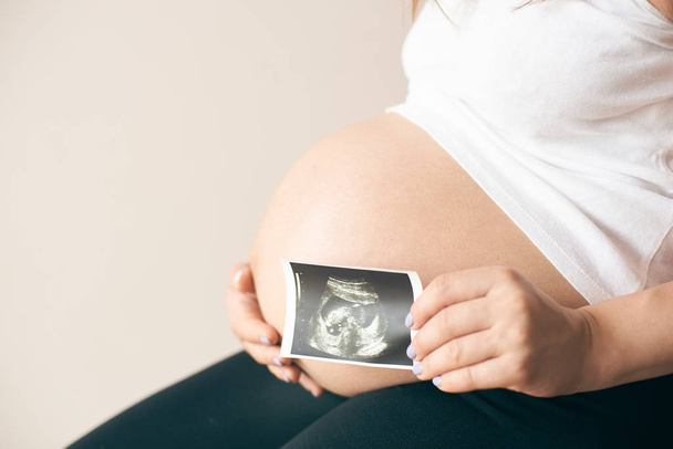 Side view of pregnant woman in white shirt sitting and showing ultrasound image of baby - Foto, imagen