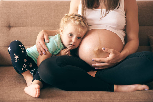 lovely girl sitting on sofa with pregnant mother and looking at camera - Foto, afbeelding