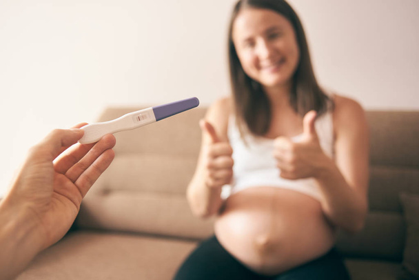 Selective focus of pregnancy test in male hands and cheerful pregnant woman at background - Photo, Image