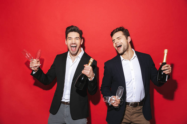 Two cheerful smartly dressed men standing isolated over red background, celebrating New Year, drinking champagne - 写真・画像