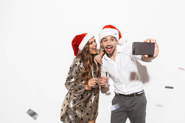 Cheerful young smartly dressed couple celebrating New Year party isolated over white background, taking a selfie, holding glasses of sparkling champagne - Foto, immagini