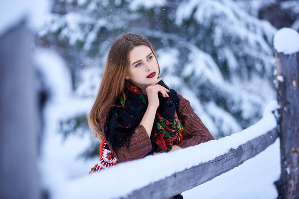 Porträt einer attraktiven jungen Frau mit langen glatten, lockeren Haaren in einem langen, stylischen Kleid aus Wolle und einem dunklen Halstuch mit hellem Blumenmuster an einem sonnigen, schneebedeckten Wintertag - Foto, Bild