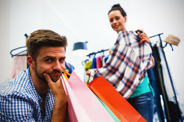 Bored man with shopping bags while woman by clothes rack at clothes store - Photo, image