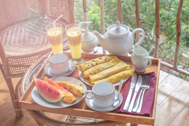 wunderschön serviertes Frühstück auf Terrasse oder Balkon, Hotelzimmer, Resort - frische Wassermelone, Mango, Banane, Smoothie, Pfannkuchen, Teekanne, Tassen, Rattanstuhl - Foto, Bild