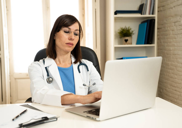 Portrait of successful attractive woman doctor in white coat working on patient medical records on laptop computer and writting on paperwork in hospital desk for Health care and Medical insurance. - Foto, Imagem