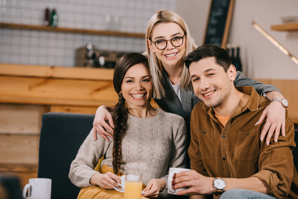 fröhliche Frau mit Brille umarmt lächelnde Freunde im Café  - Foto, Bild