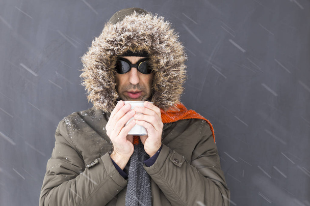 Portrait d'un aventurier d'hiver célibataire portant un manteau vert chaud avec capuche en fourrure, une écharpe orange et des lunettes de style rétro noir buvant dans une tasse en métal
 - Photo, image