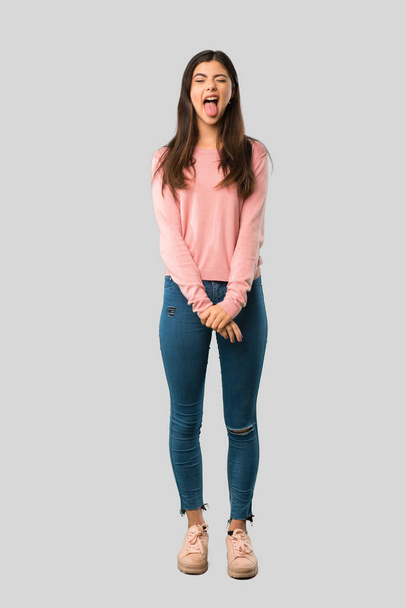 Cuerpo completo de adolescente chica con camisa rosa mostrando la lengua en la cámara que tiene mirada divertida sobre fondo gris aislado
 - Foto, Imagen