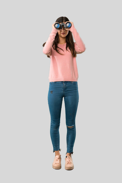 Full body of Teenager girl with pink shirt and looking in the distance with binoculars on isolated grey background - Фото, зображення