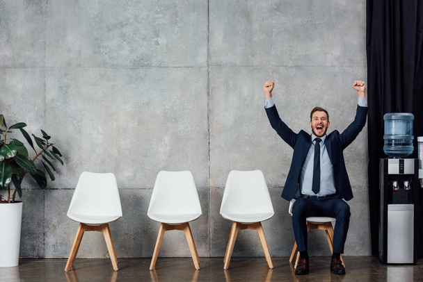 excited businessman sitting on chair and cheering with clenched fists in waiting hall - Foto, Imagem