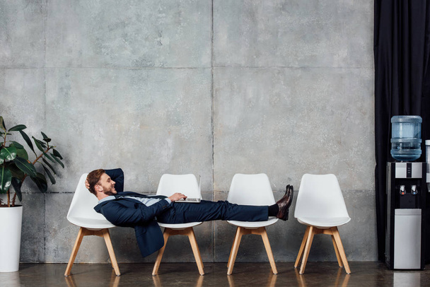 smiling businessman lying on chairs and using laptop in waiting hall - Photo, image