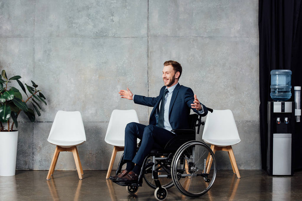 χαμογελώντας επιχειρηματίας με απλωμένα χέρια που κάθεται σε αναπηρικό καροτσάκι στην αίθουσα αναμονής  - Φωτογραφία, εικόνα