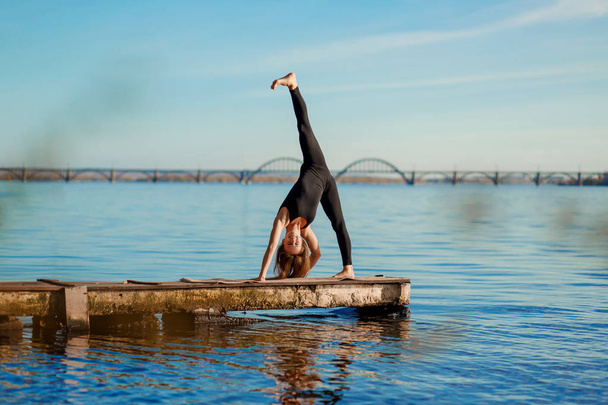 Young woman practicing yoga exercise at quiet wooden pier with city background. Sport and recreation in city rush. - Photo, Image