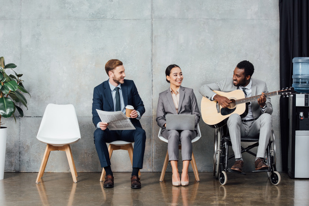 smiling multicultural businesspeople sitting and listening while african american man in wheelchair playing guitar in waiting hall - Foto, immagini