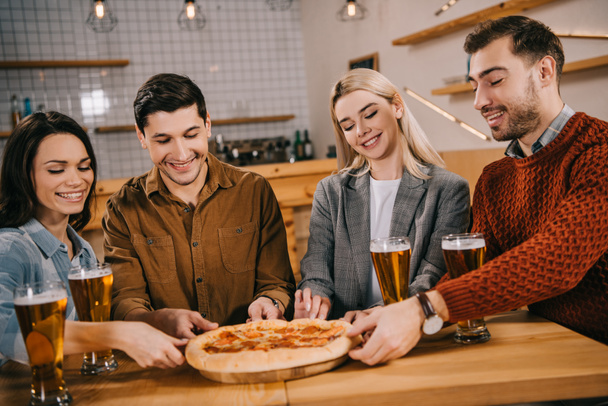 cheerful friends smiling while taking pieces of tasty pizza in bar - Fotografie, Obrázek