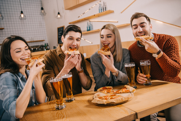happy friends eating pizza near glasses of beer in bar - Фото, изображение