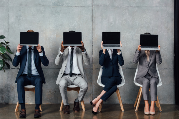 empresarios en ropa formal sentados en sillas y portátiles con pantalla en blanco frente a las caras en la sala de espera
 - Foto, imagen