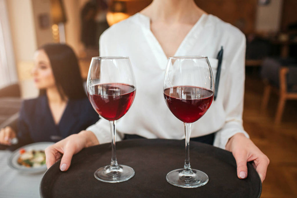 Cut view of waitress hold two glasses of red wine on tray. Youn businesswoman sit behind her at table and look left. She has salad bowl on table. - Фото, изображение