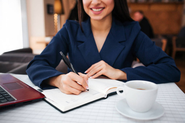 Cut view of cheerful young woman smile and write in notebook. She has laptop and cup of coffee at table. Young businesswoman work in restaurant. - Photo, Image