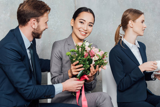 zakenman presenteren bloemen aan mooie lachende Aziatische zakenvrouw in kantoor - Foto, afbeelding