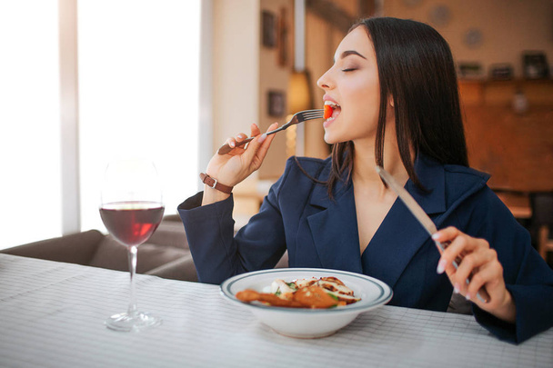 Young woman sit at table in restaurant and enjoy eating salad. She keep eyes closed. Glass of red wine stand on table. - Foto, Bild