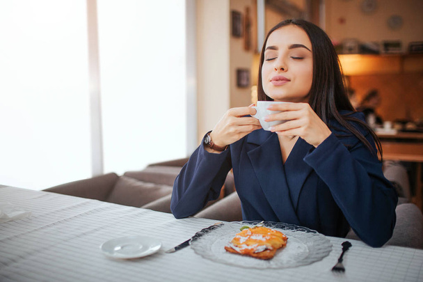 Young woman enjoying her cup of coffee. She sit at table and smell it. Model has cake on plate on table. - Foto, Imagen