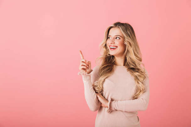 Portrait of a cheerful young woman wearing sweater standing isolated over pink background, pointing away at copy space - Zdjęcie, obraz