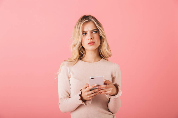 Portrait of an upset young woman wearing sweater standing isolated over pink background, using mobile phone - Fotoğraf, Görsel