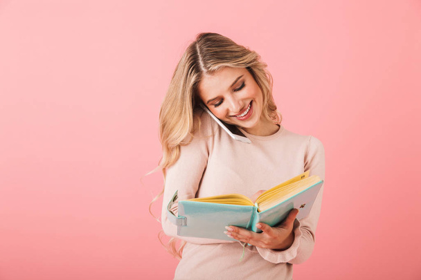 Portrait of a cheerful young woman wearing sweater standing isolated over pink background, writing notes in a diary while talking on mobile phone - Photo, image