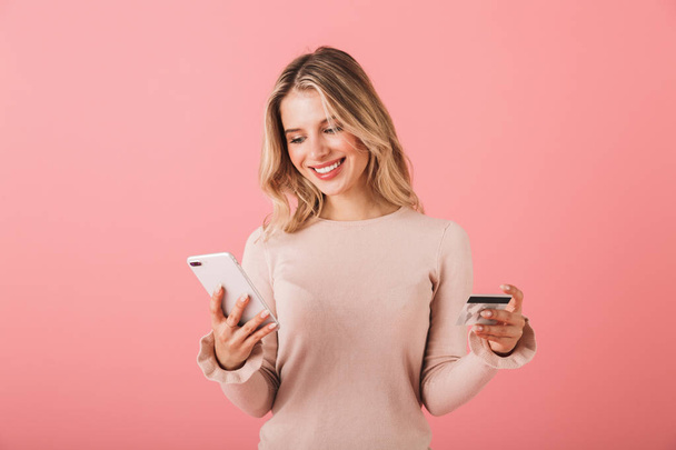 Portrait of a happy young woman wearing sweater standing isolated over pink background, using mobile phone, showing credit card - Фото, изображение
