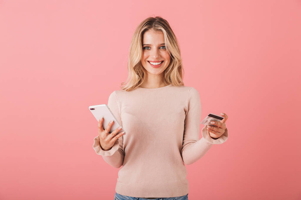 Portrait of a happy young woman wearing sweater standing isolated over pink background, using mobile phone, showing credit card - Foto, imagen