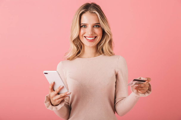Portrait of a happy young woman wearing sweater standing isolated over pink background, using mobile phone, showing credit card - 写真・画像