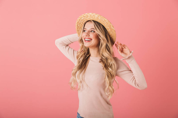 Portrait of a lovely blonde haired young woman wearing straw hat standing isolated over pink background, posing - 写真・画像