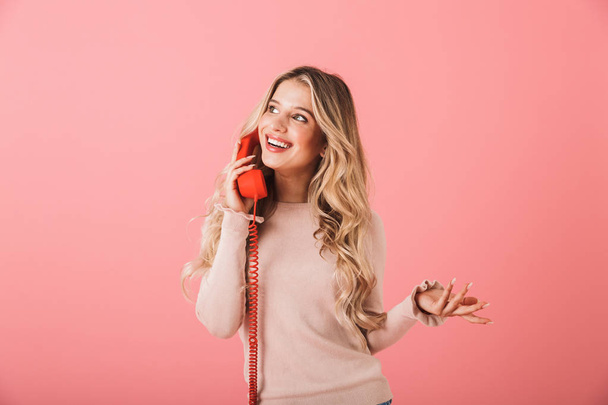 Portrait of a smiling blonde haired young woman standing isolated over pink background, talking on a landline phone - Фото, изображение