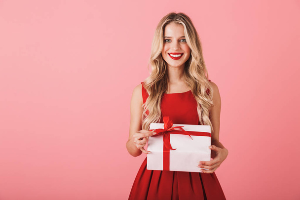 Portrait of a smiling young woman in red dress standing isolated over white background, holding present box - 写真・画像