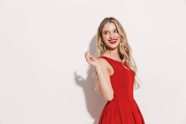 Portrait of a smiling young woman in red dress standing isolated over white background, looking away - Foto, immagini