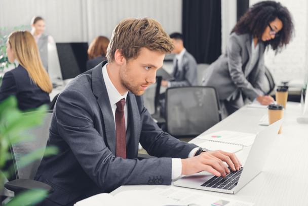 jeune homme d'affaires concentré utilisant un ordinateur portable tout en travaillant avec des collègues dans un bureau ouvert
  - Photo, image