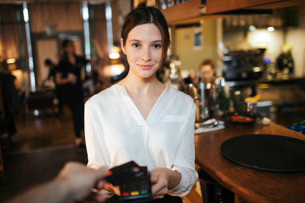 Young cheerful waitress hold paying terminal in hands and look straight on camera. Hand hold credit card upon terminal. He pays. - Φωτογραφία, εικόνα