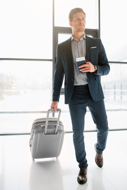 Portrait of walking young businessman walking in airport hall. He rolls suitcase and hold passport with ticket in it. Guy looks serious. - 写真・画像