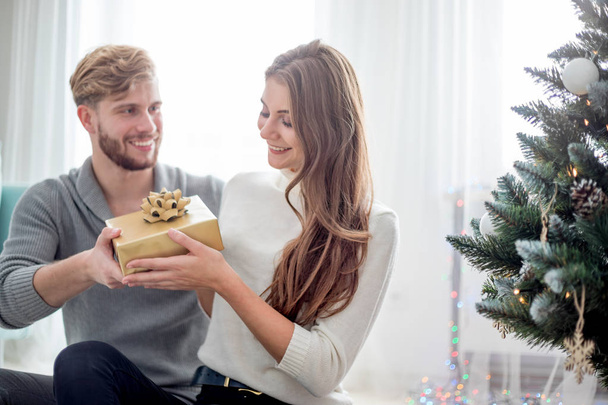Happy couple are giving themselves Christmas gifts sitting on floor next to Xmas tree - Foto, afbeelding