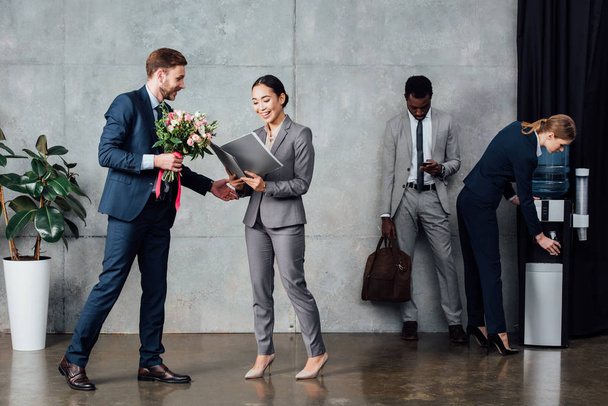 бизнесмен вручает цветы улыбающейся бизнесвумен с многонациональными коллегами на заднем плане в зале ожидания
 - Фото, изображение