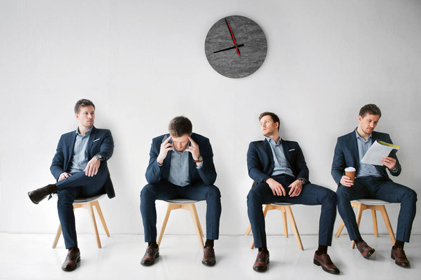 Concept waiting for a meeting. Four young man in suits sitting on white chairs in one room. First look to right. Second has headache. Third lean to wall. Fourth read menu and hold cup of coffee. - Fotoğraf, Görsel