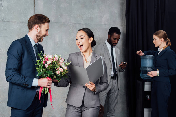 hombre de negocios presentando flores a alegre mujer de negocios asiática con colegas en fondo en sala de espera
 - Foto, imagen